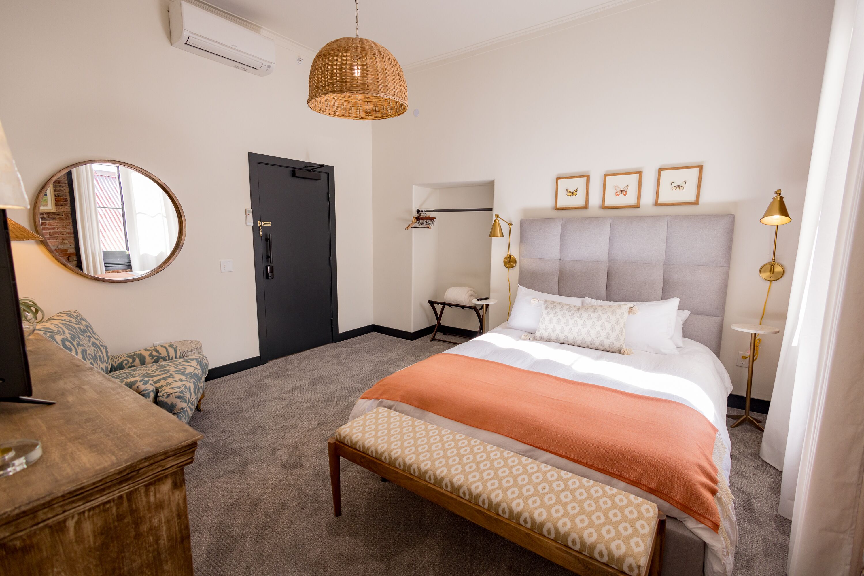 A hotel room with a bed, a bench, a desk with a lamp, a mirror, and a wall-mounted air conditioner.