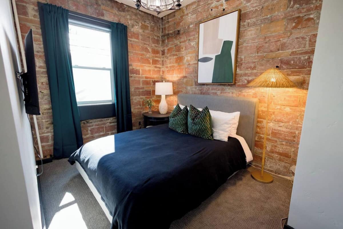 Quarto com parede de tijolos, cama com cobertura escura, luminária e um quadro na parede.