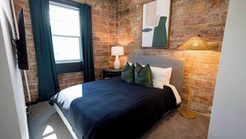 Un dormitorio con pared de ladrillo, una cama con cubierta oscura, una lámpara y un cuadro en la pared.