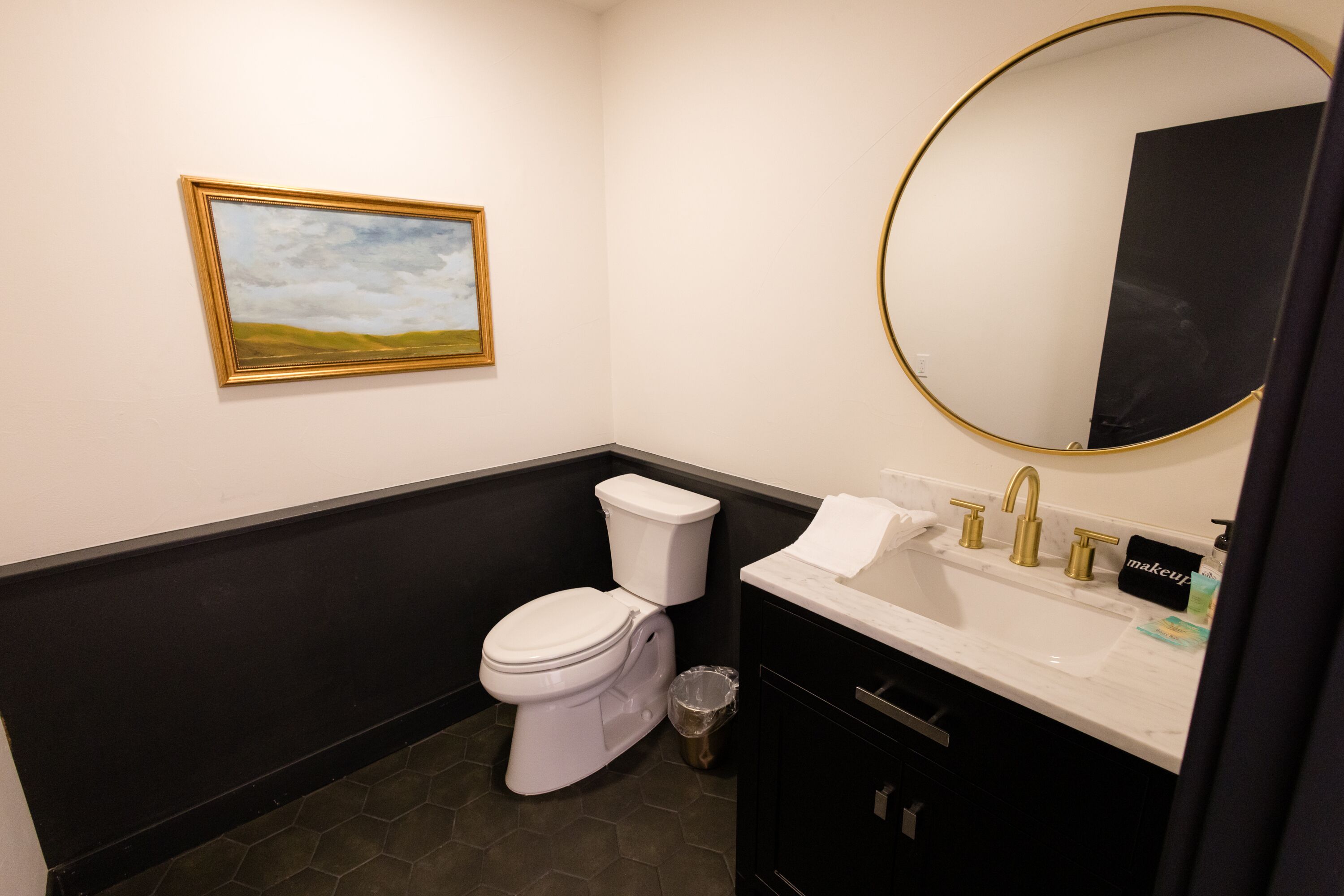 A bathroom with a white toilet, a sink with gold fixtures, a black vanity, a round mirror, and a framed landscape painting on the wall.