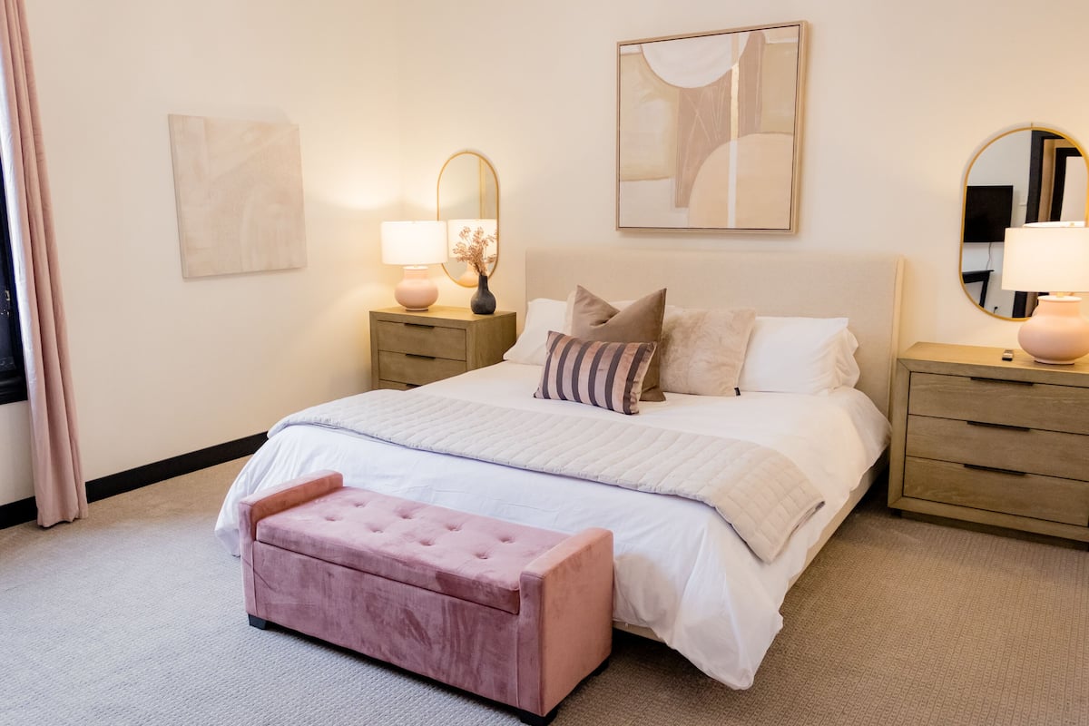 Quarto bem arrumado com uma cama grande, mesas de cabeceira, um banquinho rosa e obras de arte na parede.
