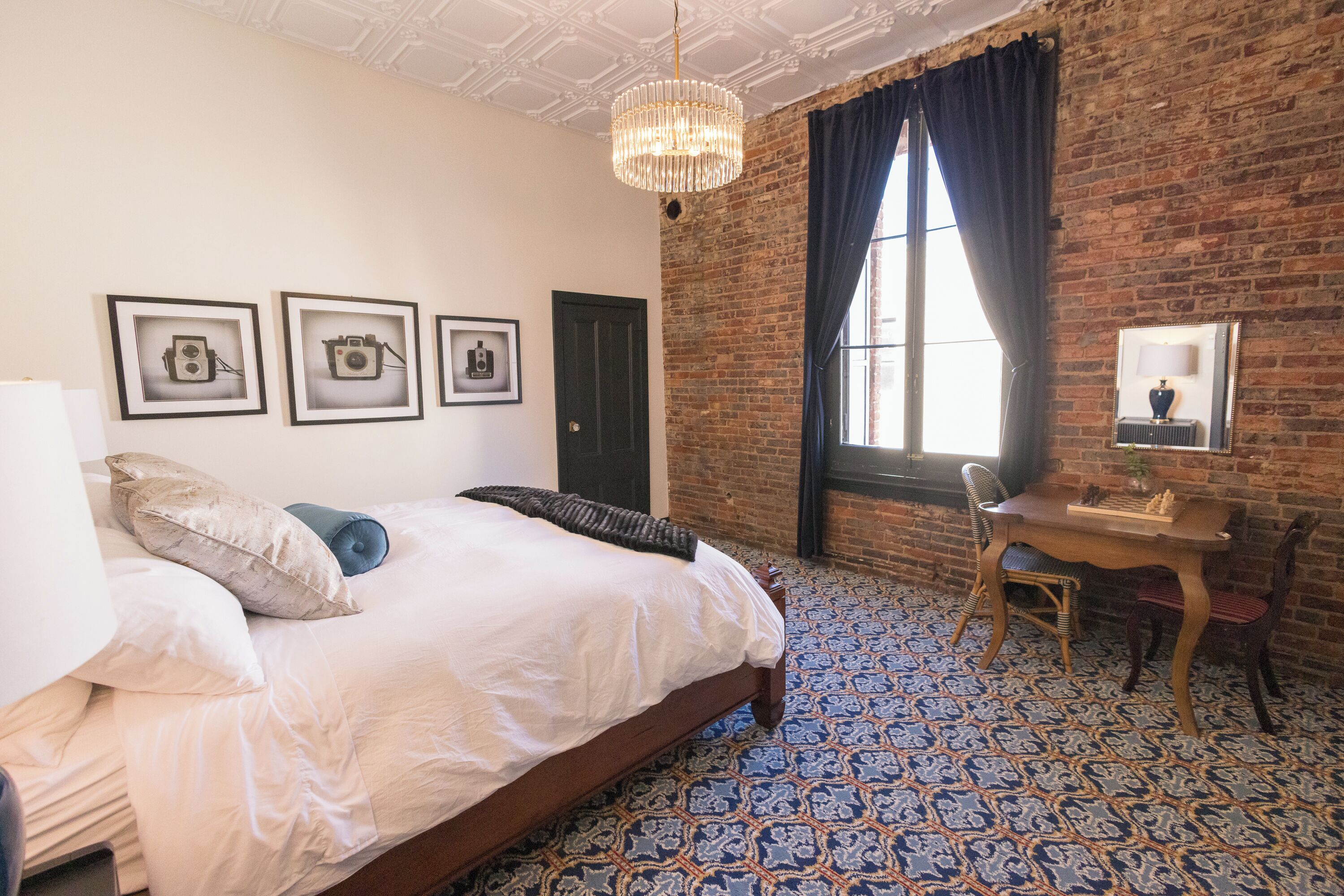A hotel room with a large bed, a desk, and a brick wall. There are three framed pictures on the wall, a chandelier, and a window with curtains.