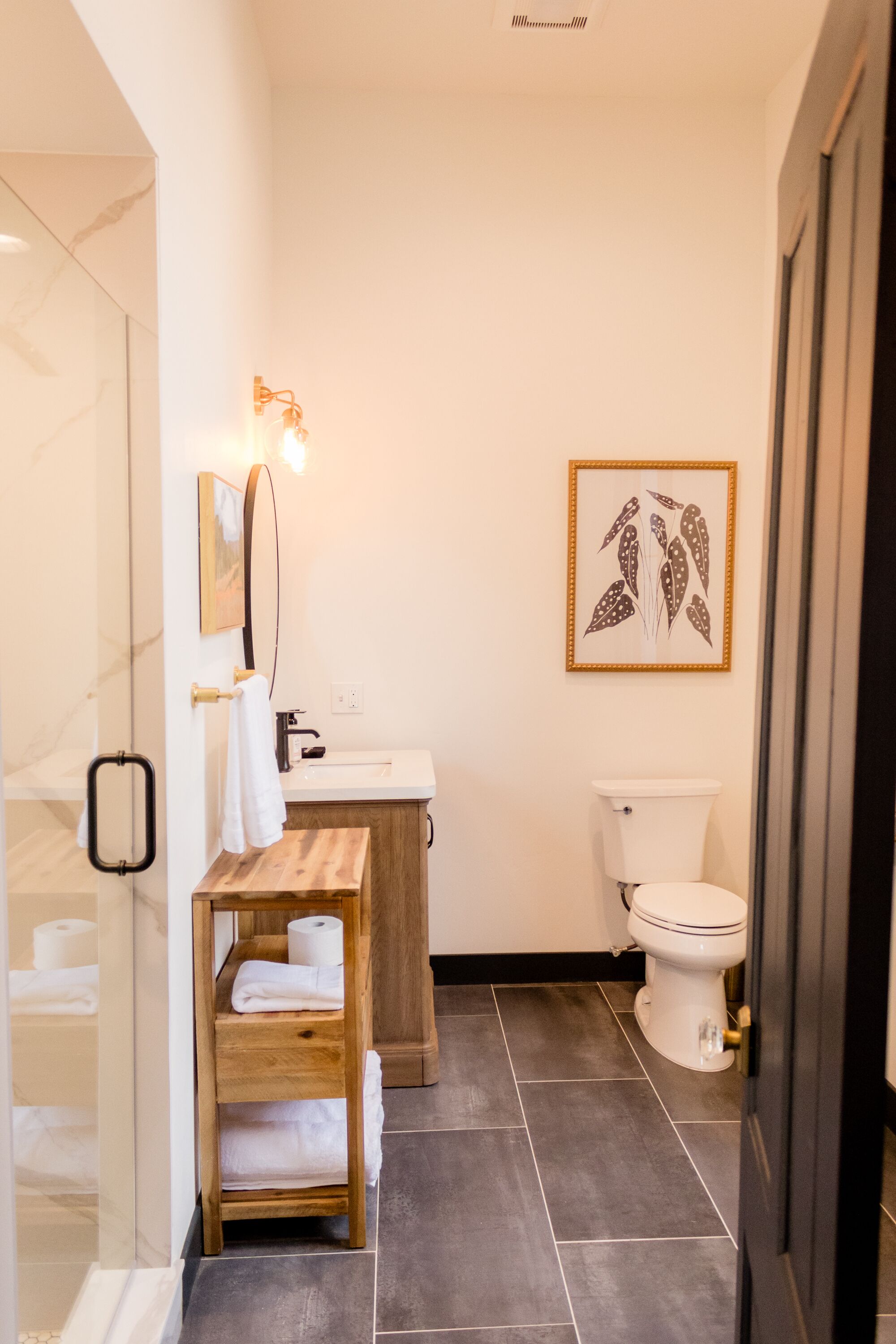 A modern bathroom with a wooden vanity, white sink, and toilet. There is a framed artwork on the wall and a towel rack with towels.