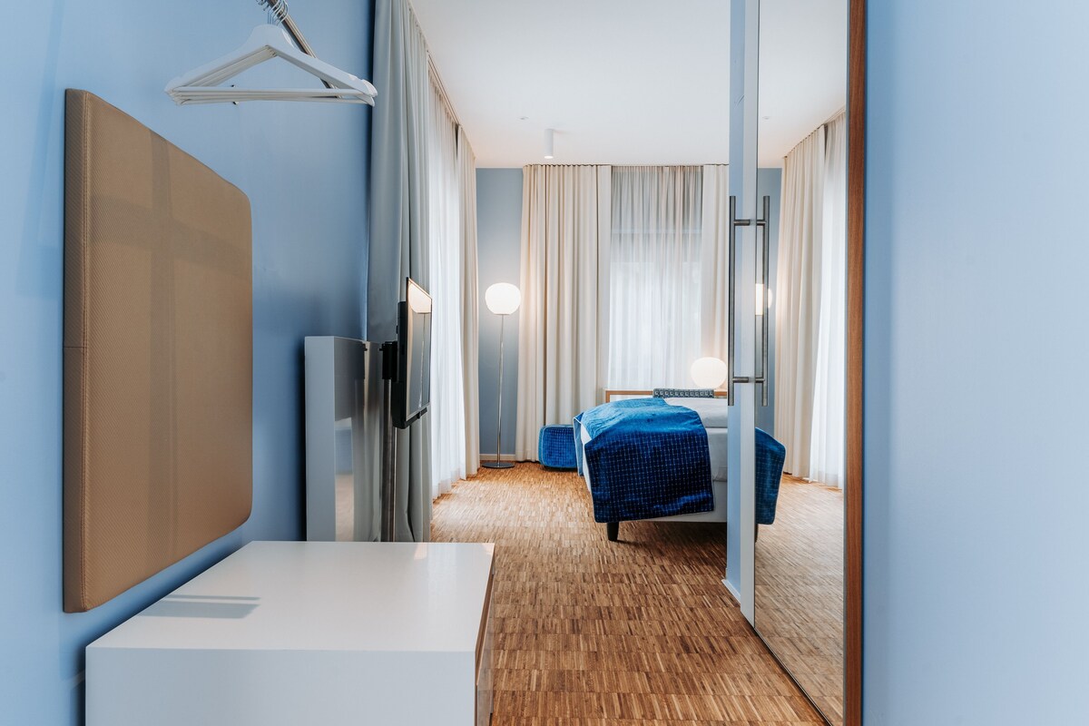 A hotel room with a bed, a desk, a chair, a mirror, and a hanging coat rack.