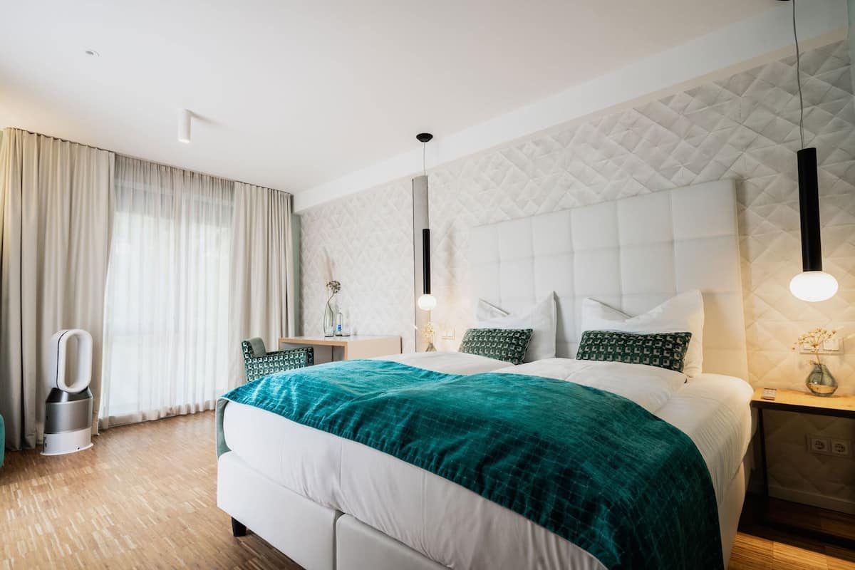 A modern hotel room with a large bed, a desk, a chair, and a bedside table.