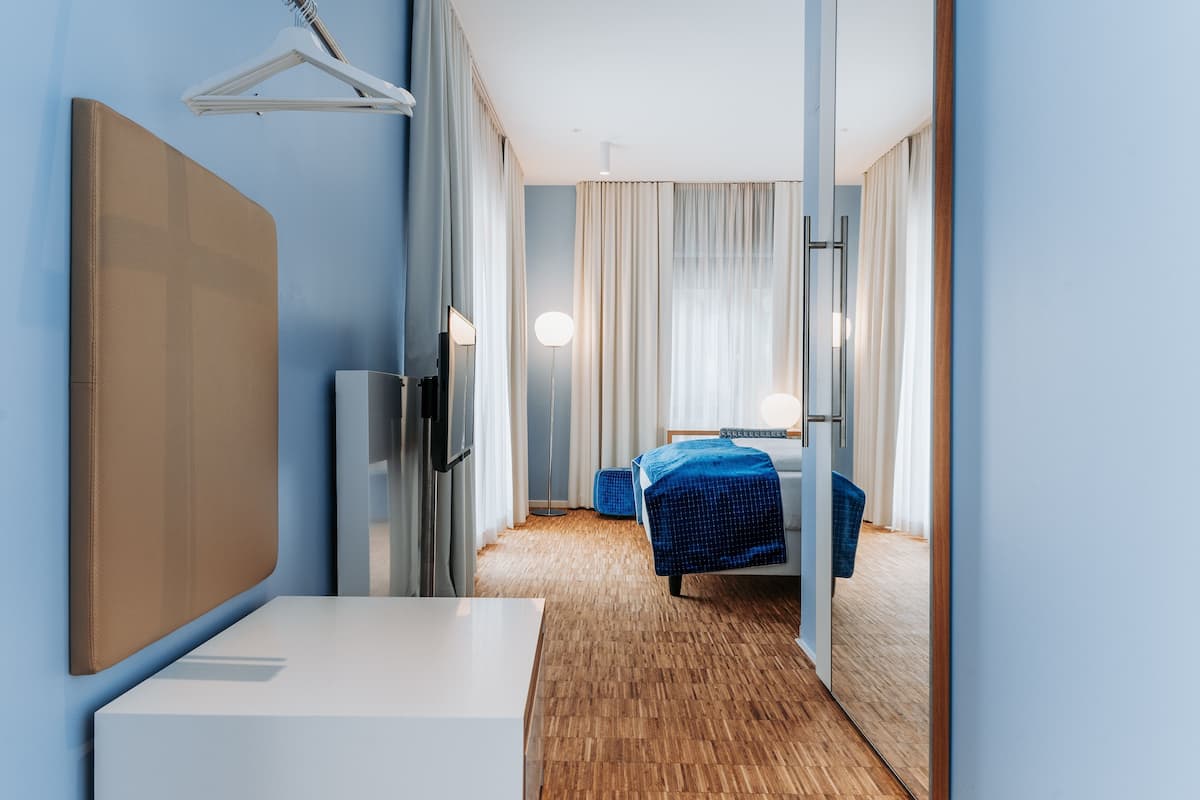 A hotel room with a bed, a desk, a chair, a mirror, and a hanging coat rack.