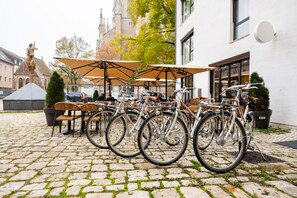 Bicicletas estacionadas sob um toldo, um pátio de paralelepípedos e um edifício com torre do relógio ao fundo.