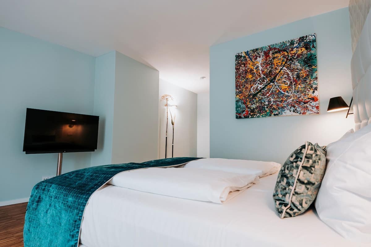 A modern hotel room with a bed, a flat-screen TV, a painting on the wall, and a floor lamp.