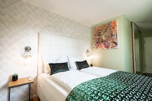 Habitació d'hotel amb sostre amb rellenu, llit amb roba de llit blanca, manta estampada de color verd i un quadre abstracte i colorit a la paret.