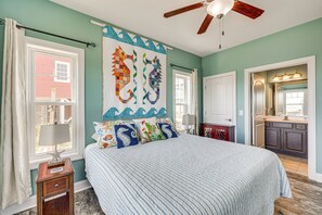 Une chambre à coucher avec un lit, une table de chevet, une fenêtre avec des rideaux, un ventilateur de plafond et une salle de bain visible à travers une porte ouverte.