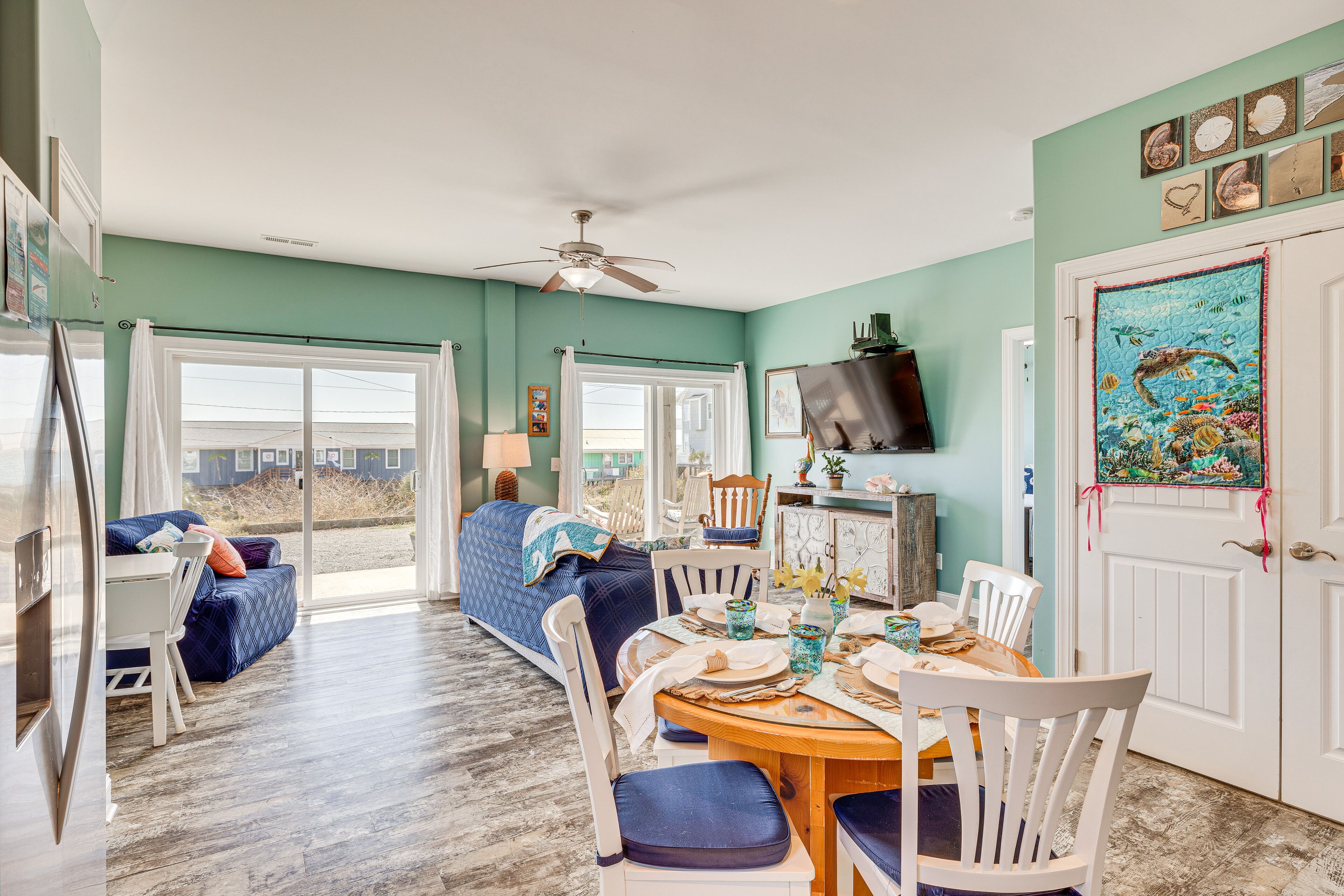 Un coin repas avec une table en bois dressée pour quatre personnes, des chaises blanches et un ventilateur de plafond.