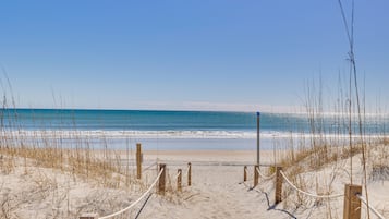 A sandy beach with a wooden walkway leading to the ocean, surrounded by tall grasses.
