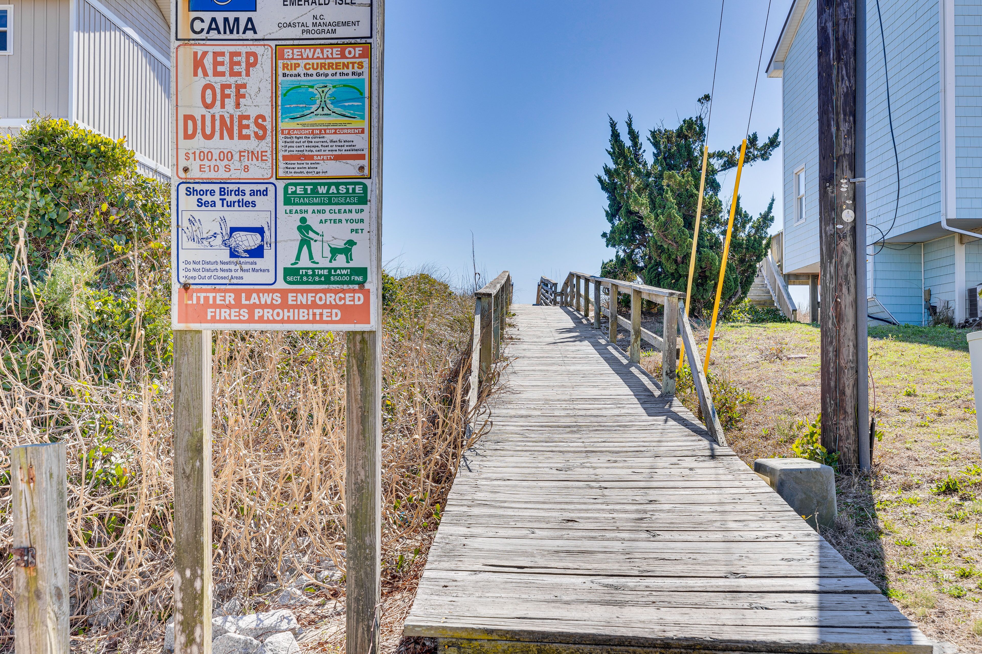 A wooden boardwalk leading to a house with multiple signs, including one about keeping off dunes and a fine for littering.