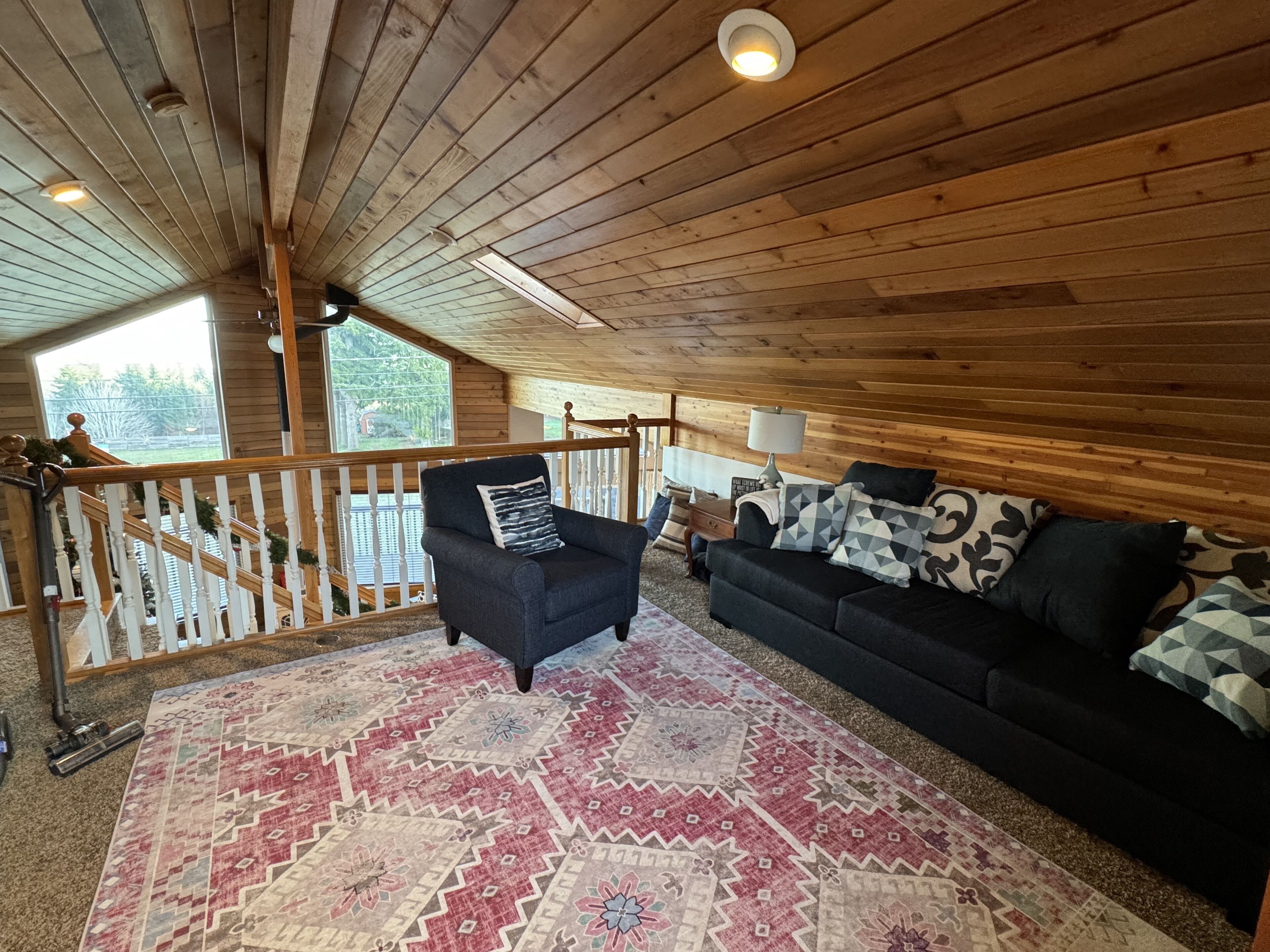 A cozy room with a wooden ceiling, a black sofa, a patterned rug, and a view of the outdoors through a window.