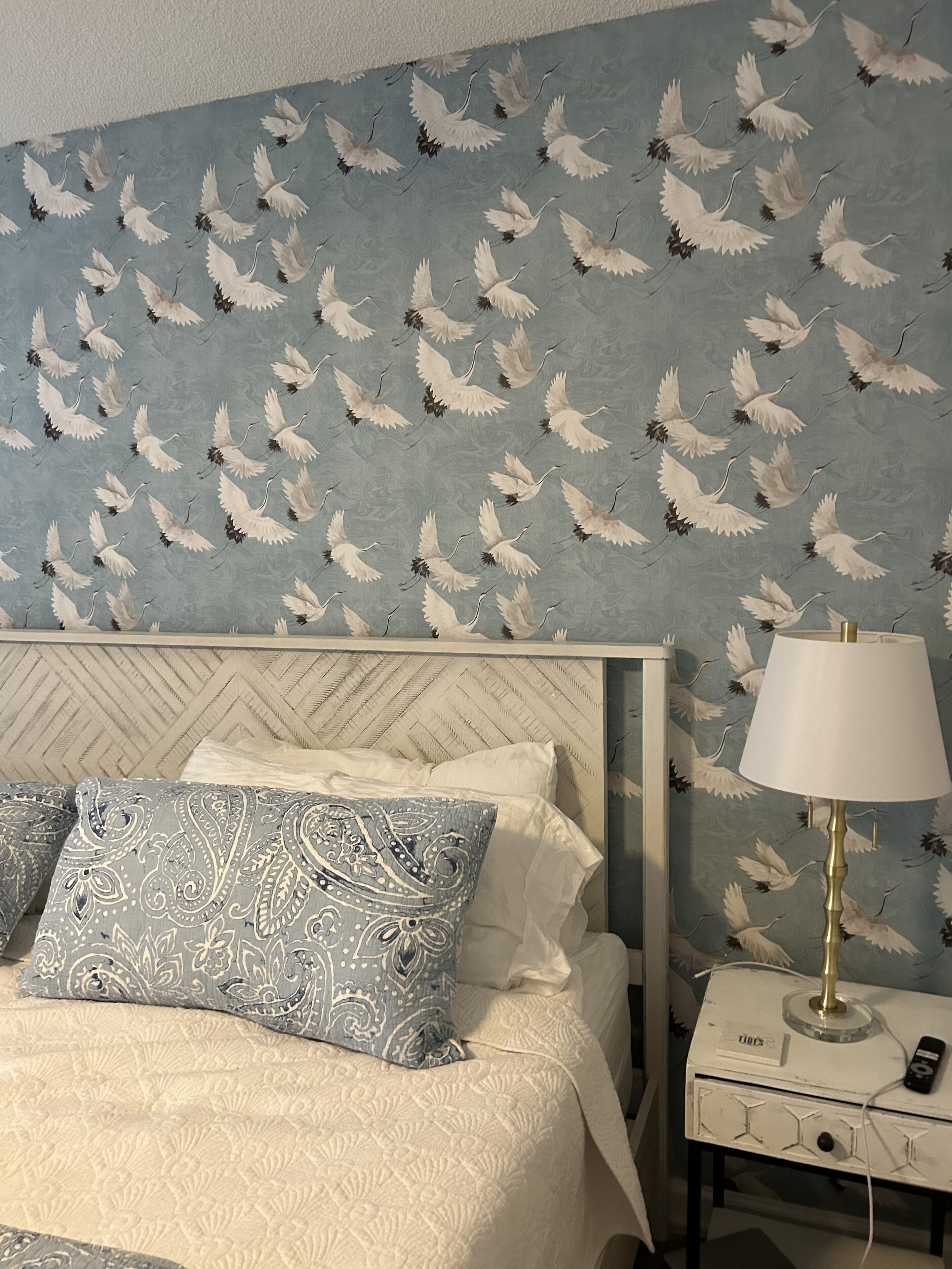 A hotel room with a patterned wallpaper, a bed with two pillows, a nightstand with a lamp, and a remote control.