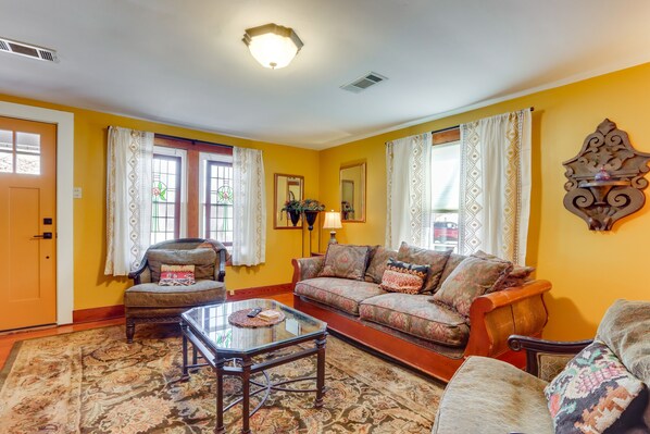 A living room with a sofa, armchair, and coffee table. There is a door, windows with curtains, and a wall-mounted decorative piece. - 2 Mi to UA: Central Fayetteville Home w/ Patios (Fayetteville)