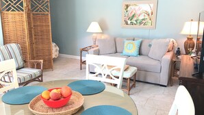 A living room with a sofa, chairs, a dining table with placemats, a fruit bowl, and a wall with a painting.