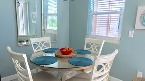 A dining area with a round glass table, white chairs, and a red bowl with fruits.