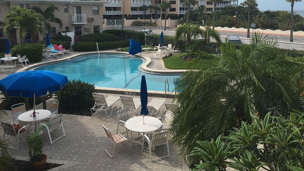 A pool area with lounge chairs and umbrellas, surrounded by greenery and a multi-story building in the background.