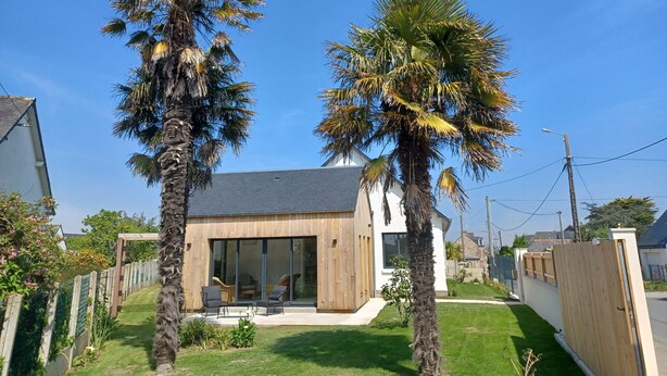Une maison moderne dotée d’une grande porte vitrée, d’une terrasse en bois et de deux palmiers dans le jardin.