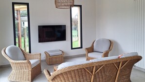 Un salon moderne avec des meubles en osier, un téléviseur à écran plat et de grandes fenêtres donnant sur un paysage verdoyant.