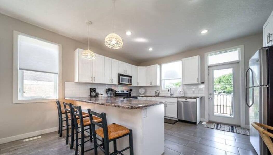 A modern kitchen with white cabinets, a central island with bar stools, a stainless steel refrigerator, and pendant lighting.