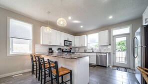 A modern kitchen with white cabinets, a central island with bar stools, a stainless steel refrigerator, and pendant lighting.
