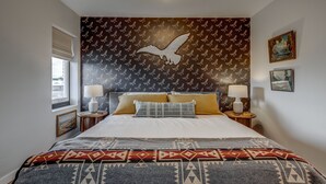 A bedroom with a large bed, patterned wallpaper, two bedside lamps, and a window with a view.