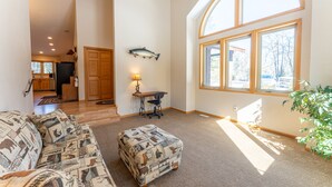 A spacious living area with a large window, a sofa with patterned cushions, a small ottoman, and a fish mounted on the wall. - Wooded setting, close to town, clean! ATV & snowmobile from front door! (Ely)