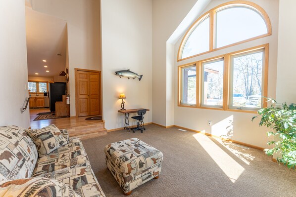 A spacious living area with a large window, a sofa with patterned cushions, a small ottoman, and a fish mounted on the wall. - Wooded setting, close to town, clean! ATV & snowmobile from front door! (Ely)