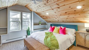 A bedroom with a sloped ceiling, a bed with colorful pillows, a bedside table, and a window with a view of trees.