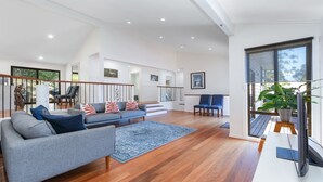 A spacious living area with a grey sofa, blue and red patterned cushions, a blue rug, and a wooden floor. There are two blue chairs, a staircase, and a balcony with a potted plant.