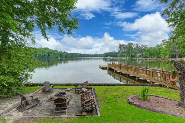 A lakeside deck with Adirondack chairs, a fire pit, and a wooden dock extending into the lake.