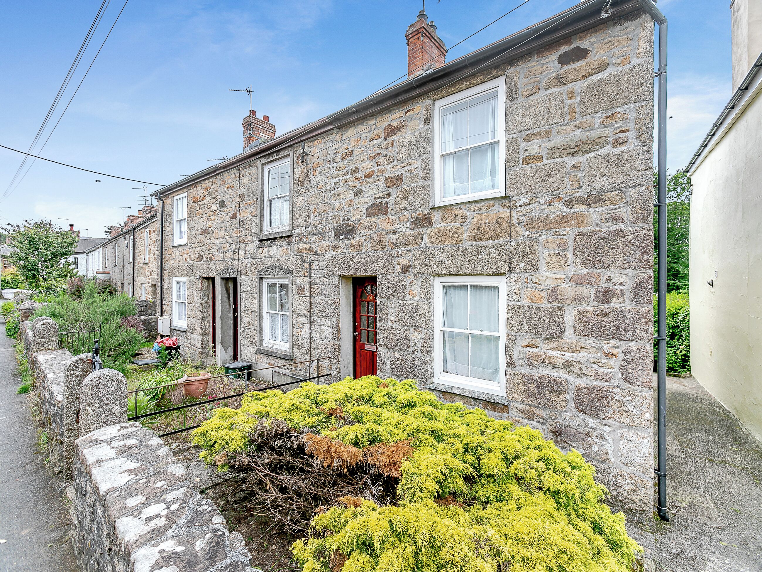 A stone house with a red door, white windows, and a small garden with yellow plants.