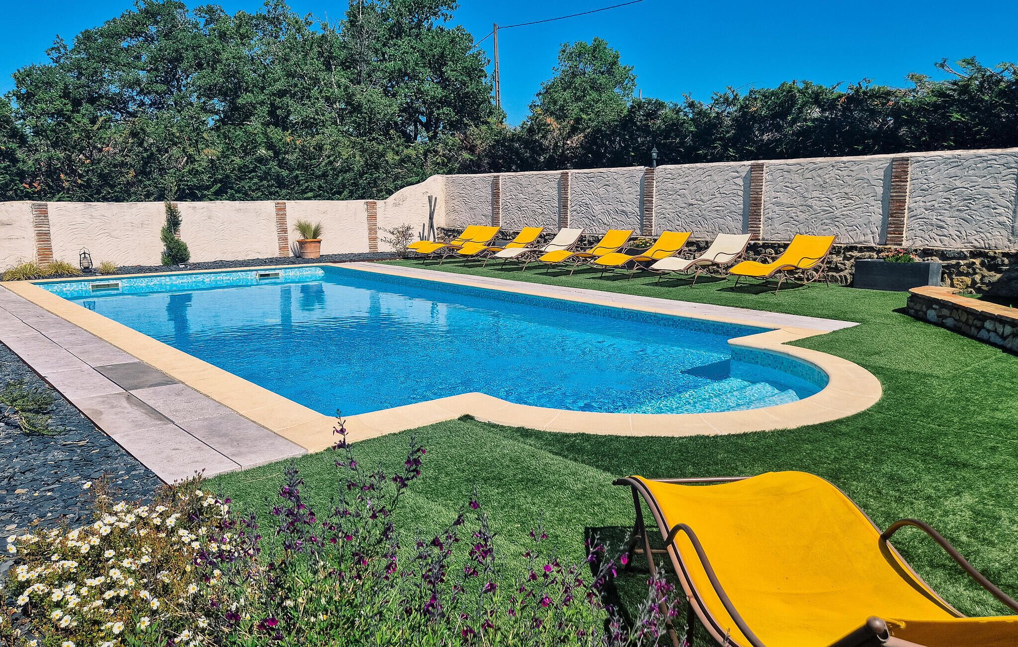 Una piscina rectangular amb un videtell daurat, envoltada per un mur de pedra i una zona enjardinada amb gandules grogues.
