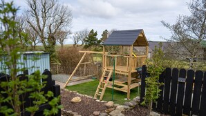 Une structure de jeu en bois, dotée d’une échelle d’escalade et d’un petit espace clos, entourée d’une clôture et d’un jardin agrémenté de paillis et de rochers.