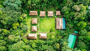 An aerial view of a resort with thatched-roof huts arranged in a square pattern, surrounded by dense tropical vegetation.