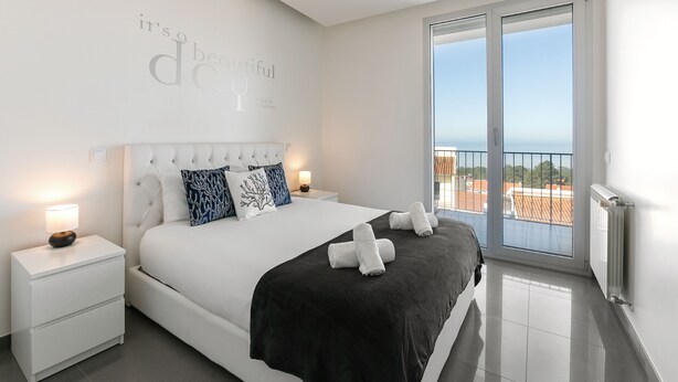 A modern bedroom with a large bed, a nightstand, a wall with a motivational quote, and a balcony with a view. - Nazaré Rooftop Oásis (Nazaré)