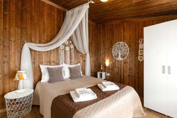 A bedroom with a wooden paneled wall, a bed with a beige quilt, a canopy, a bedside table with a lamp, and a wardrobe.