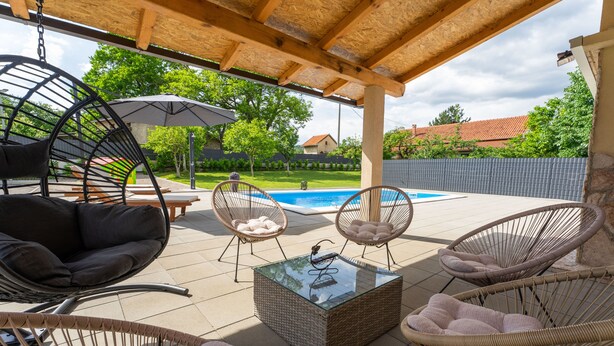 En overdækket terrasse med fletningsmøbler, et glasbord og udsigt til en swimmingpool.