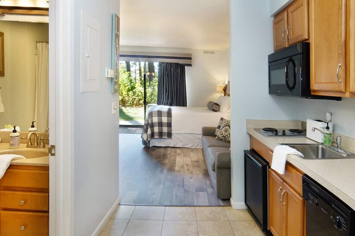 Cocina compacta con microondas, estufa y lavamanos. Se ve una cama con una colcha de cuadros a través de una puerta abierta.