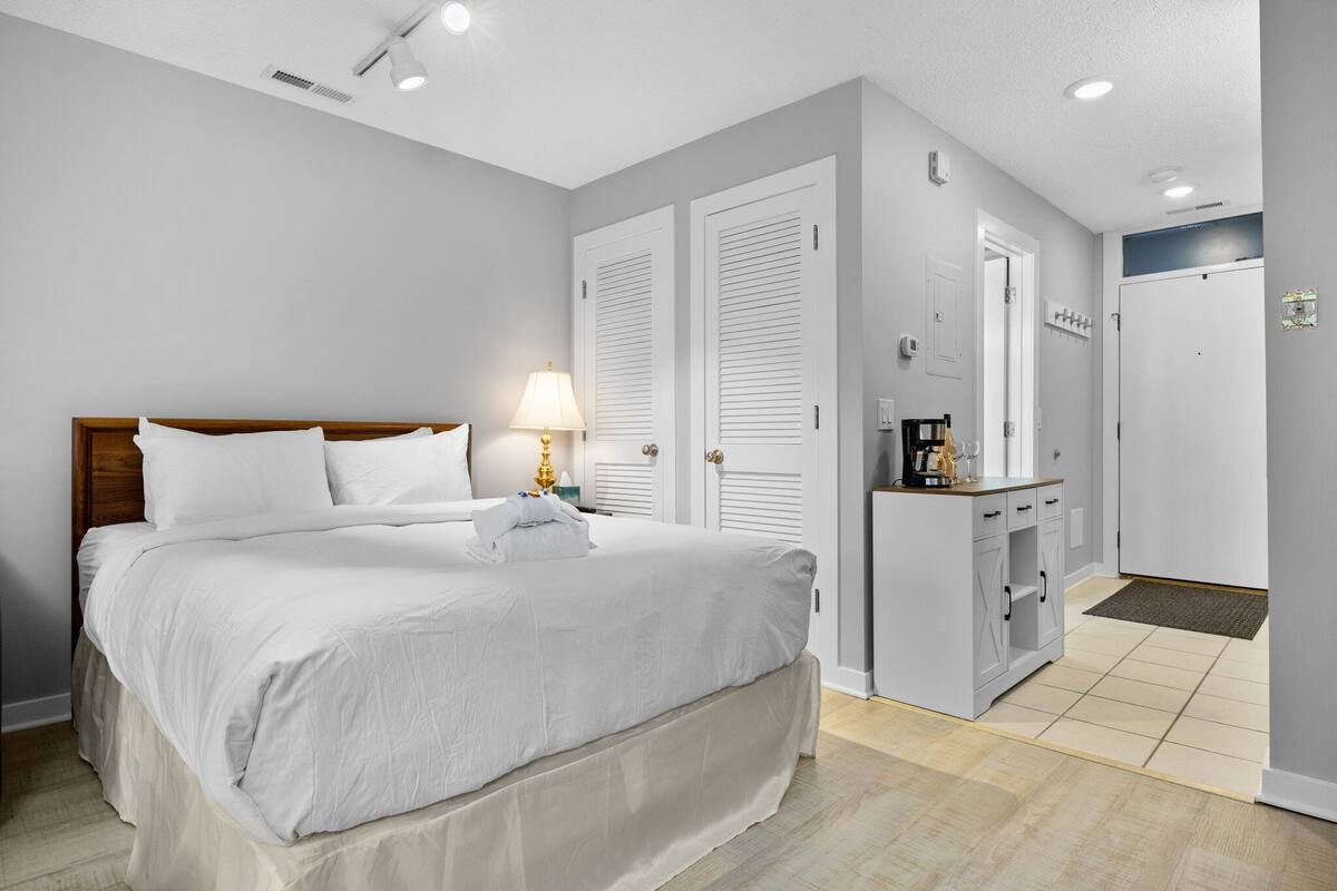Un dormitorio ordenado con cama, lámpara de noche y una mesita con cafetera.