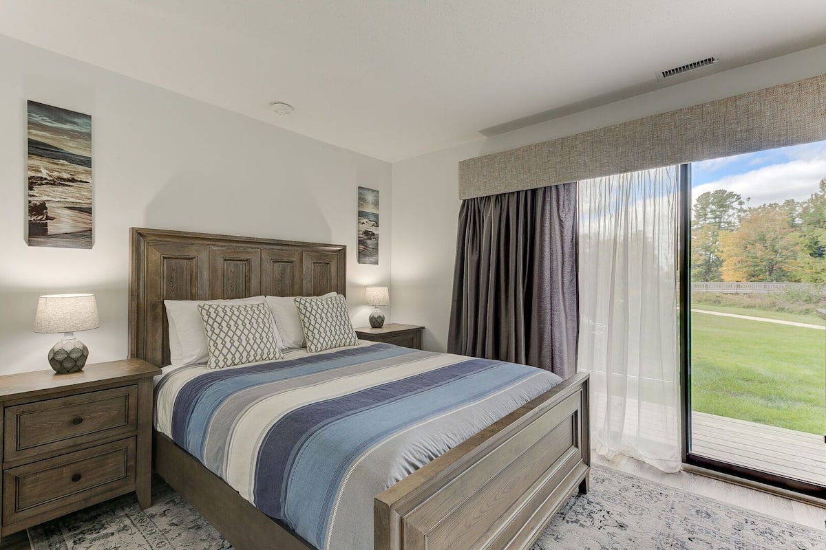 Un dormitorio con una cama grande, dos mesitas de noche y vistas a una zona verde a través de una ventana con cortinas.