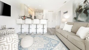 En moderne stue med en beige sofa, en mønstret skammel og et blåt og hvidt tæppe. Fire hvide barstole og et fjernsyn monteret på væggen.