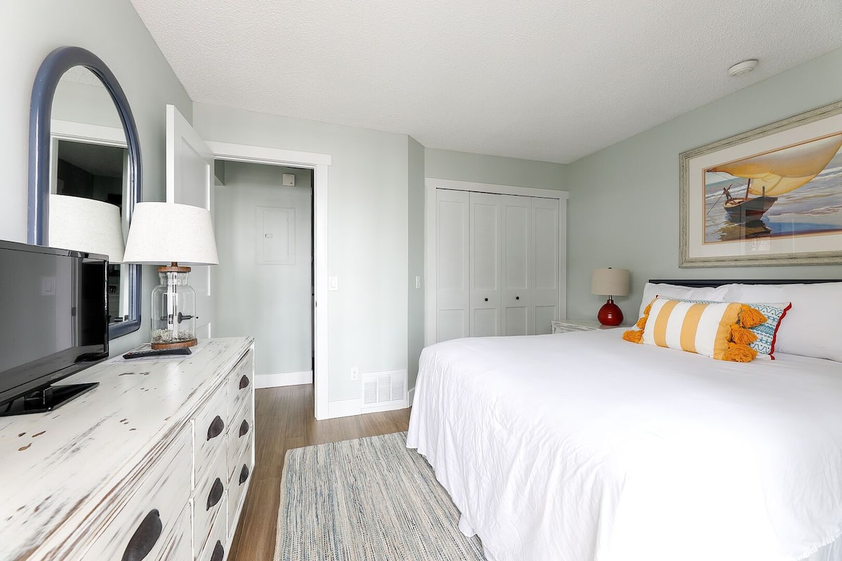 Un dormitorio con cama, cómoda, televisor y puerta que da a otra habitación.