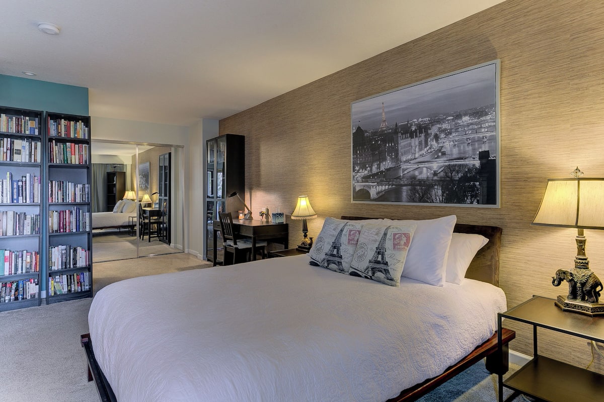 Habitación de hotel con una cama grande, una estantería, un escritorio y un cuadro de gran formato con una vista de la ciudad.