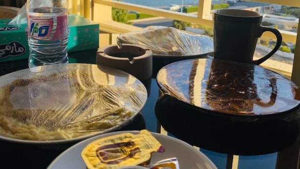 A table with a bottle of water, a mug, a plate with food, and a cup of coffee, with a view of a cityscape through a window.