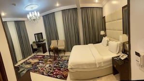 A hotel room with a large bed, a desk with a chair, a desk lamp, a telephone, and a patterned carpet.