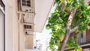 Uma varanda estreita com um banco de madeira, uma pequena mesa com um vaso de planta e um copo de vinho tinto, e vista para a rua e prédios.