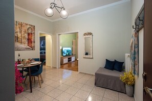Un área de comedor con una mesa dispuesta para dos personas, un sofá, un televisor al fondo y un espejo en la pared.